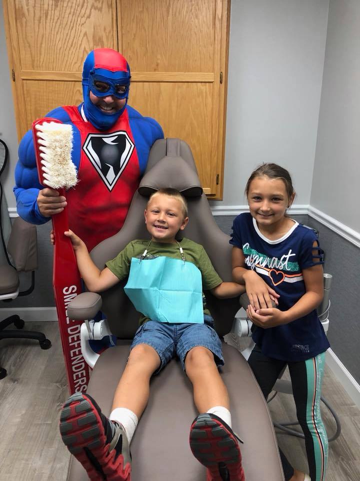 Photo of Captain Super Smiles standing next to two young dental patients who are sitting in the dental chair.
