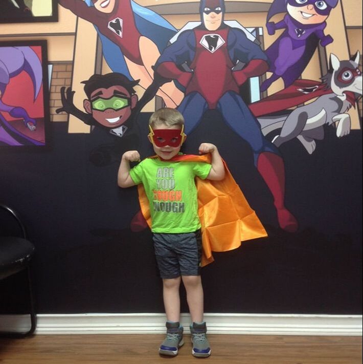 Picture of a young boy wearing a smile defender cape and mask - making muscles and standing in a super hero pose.